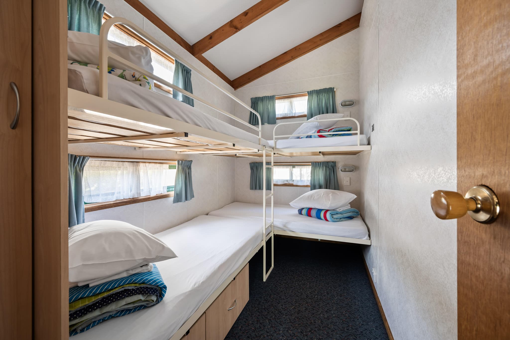 Four-bed bunk room with white metal bunks, exposed timber beams and teal curtains at Bright Accommodation Park
